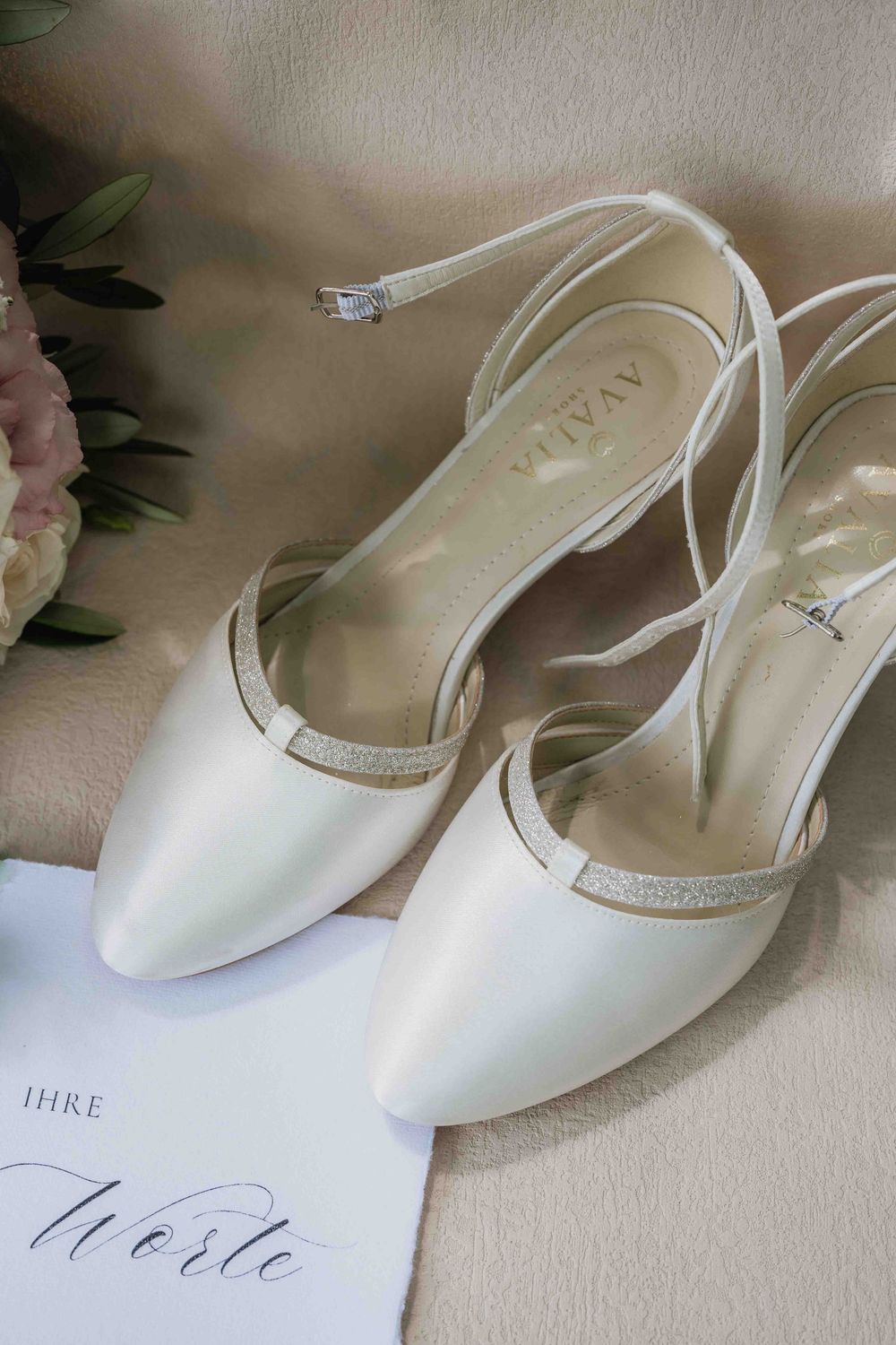 White pointed-toe bridal flats with delicate ankle straps displayed on a table with wedding stationery.