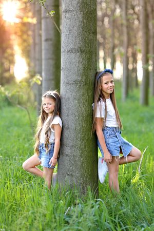 Two children enjoying a summer day leaning against a tree trunk in a sunlit forest setting.