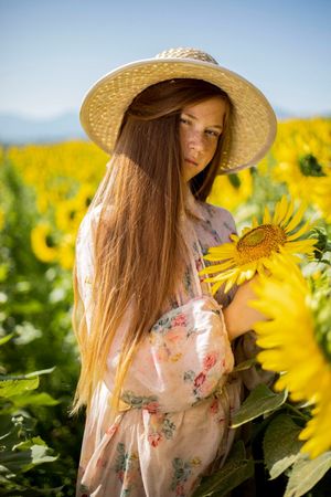 Person in straw hat and floral dress admiring sunflower in bright yellow field.