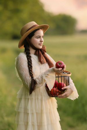 A vintage-inspired scene showing a white lace dress and woven basket filled with red apples in a sunlit meadow.