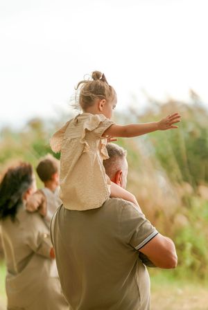 A young girl sits on a man's shoulders outdoors, arms outstretched, as a family walks through a sunny field.