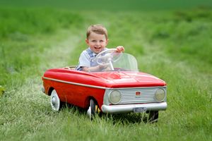 Child riding red vintage pedal car on green grass lawn.