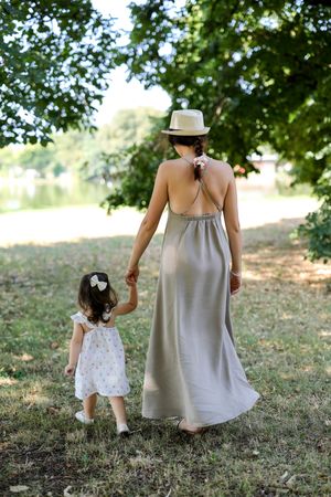 A mother in a gray maxi dress and hat holds her toddler daughter's hand while walking in a lush green park.