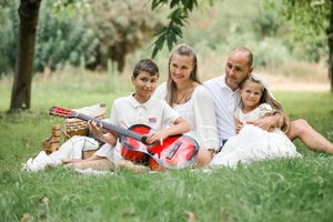 Family of four enjoying a picnic in the park, with a boy playing a red guitar on a sunny day.