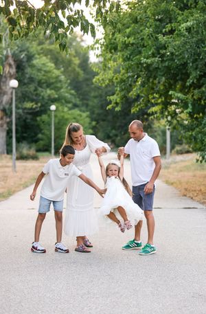 Happy family of four swinging little girl in white dress on sunny park path surrounded by green trees.