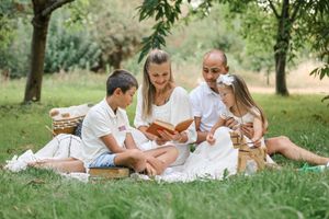 Family of four enjoying a sunny outdoor picnic on grass, reading a book together surrounded by trees.