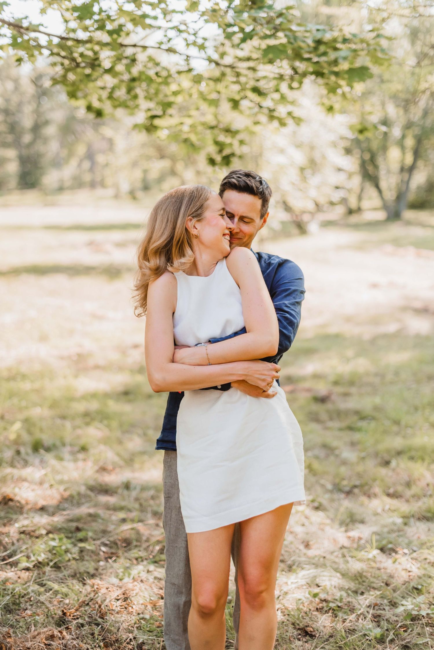 A couple embraces tenderly in a sunlit outdoor setting with trees and fallen leaves in the background.