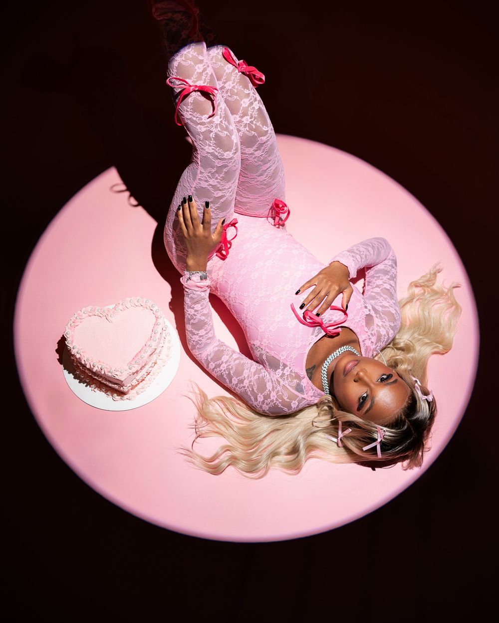 Fashion photo of a model in pink lace outfit lying on a pink circular platform with a heart-shaped prop.