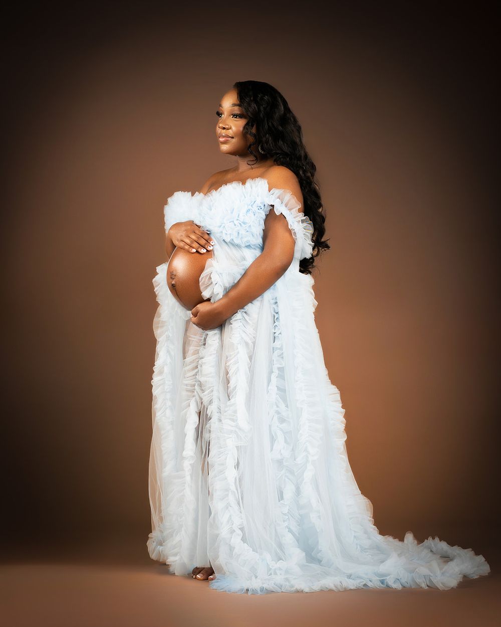 Elegant maternity photo featuring a flowing white off-shoulder gown against brown backdrop.