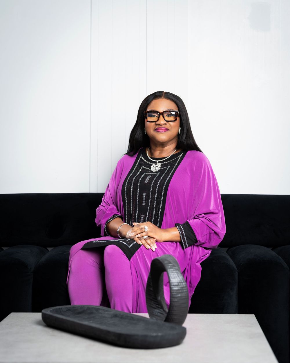 Relaxed poses on black couch wearing vibrant purple outfit and glasses.