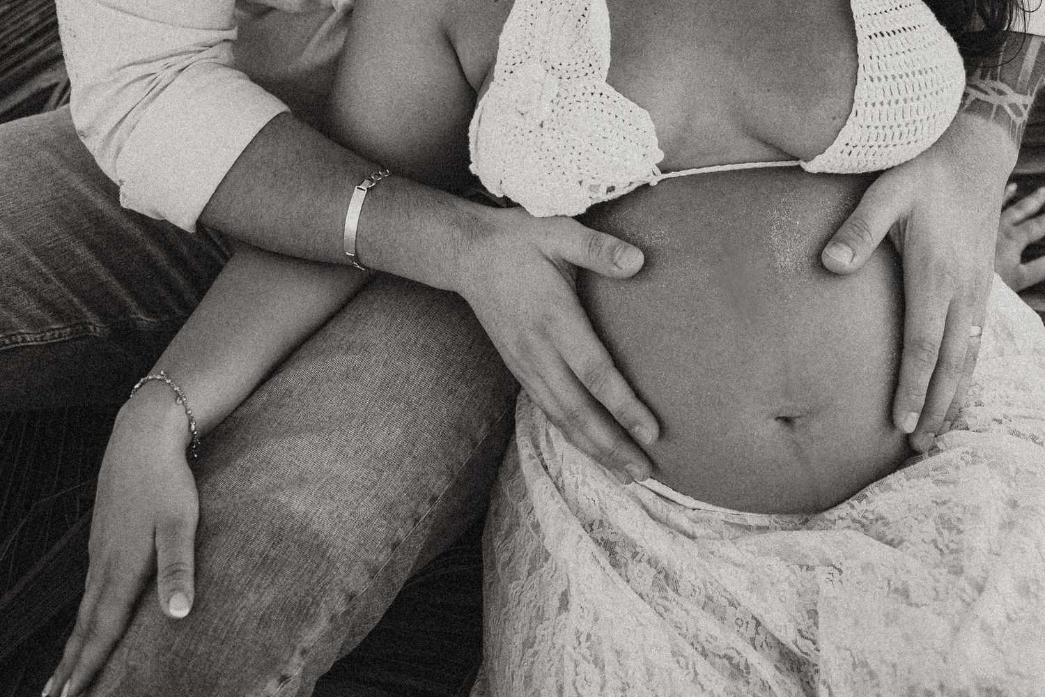 Close-up black and white shot of hands holding a heart shape against a pregnant belly.