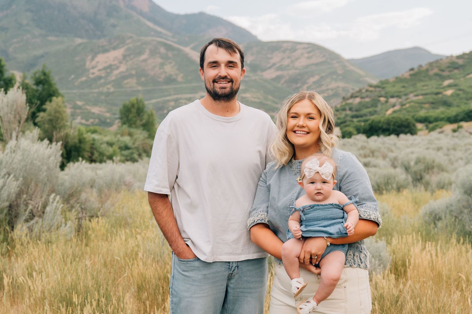 A family stands together in casual clothing against a backdrop of mountains and dry grasses in a rural landscape.