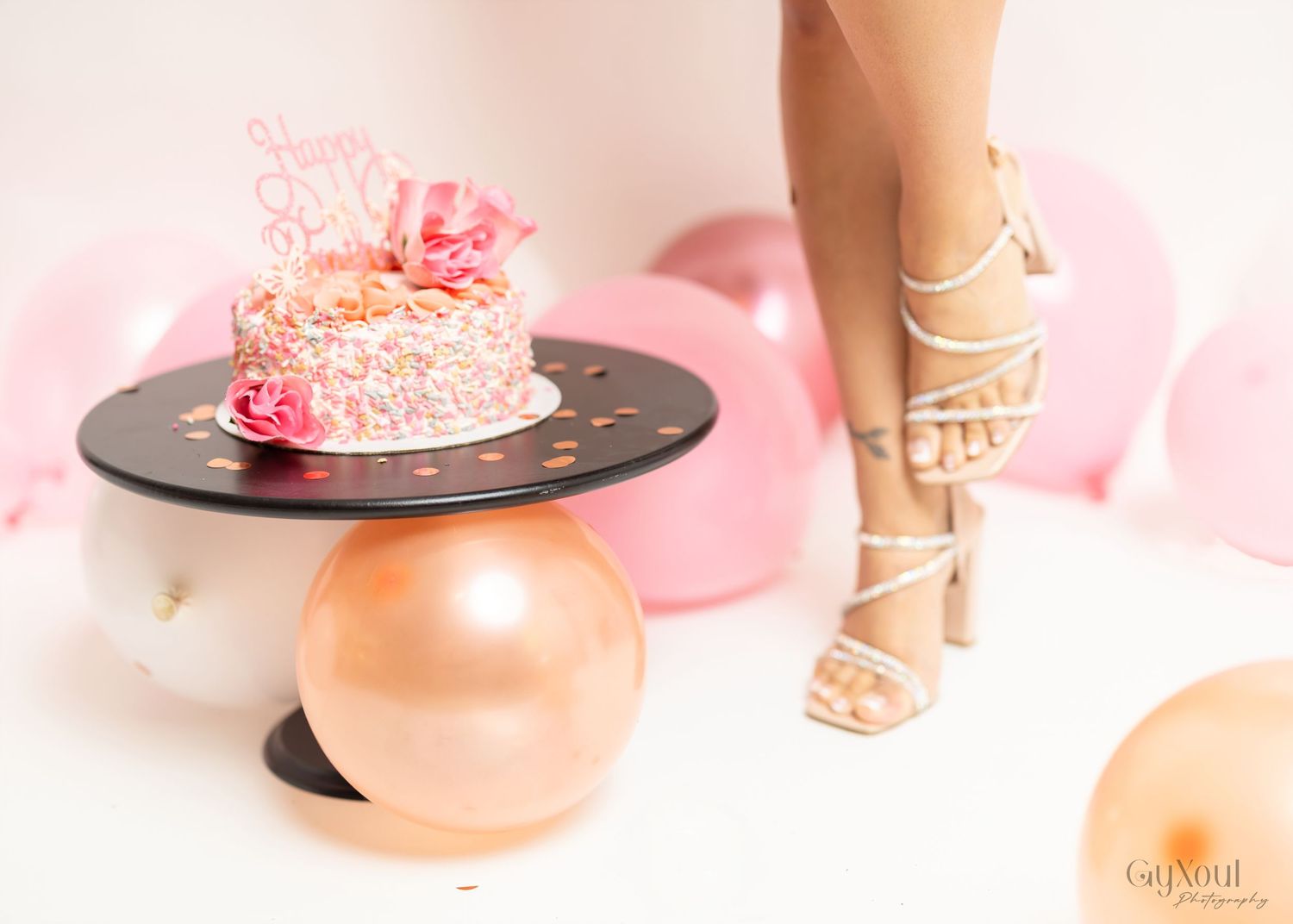 Pink birthday cake on black plate with strappy heels and rose gold balloons in feminine party setting.