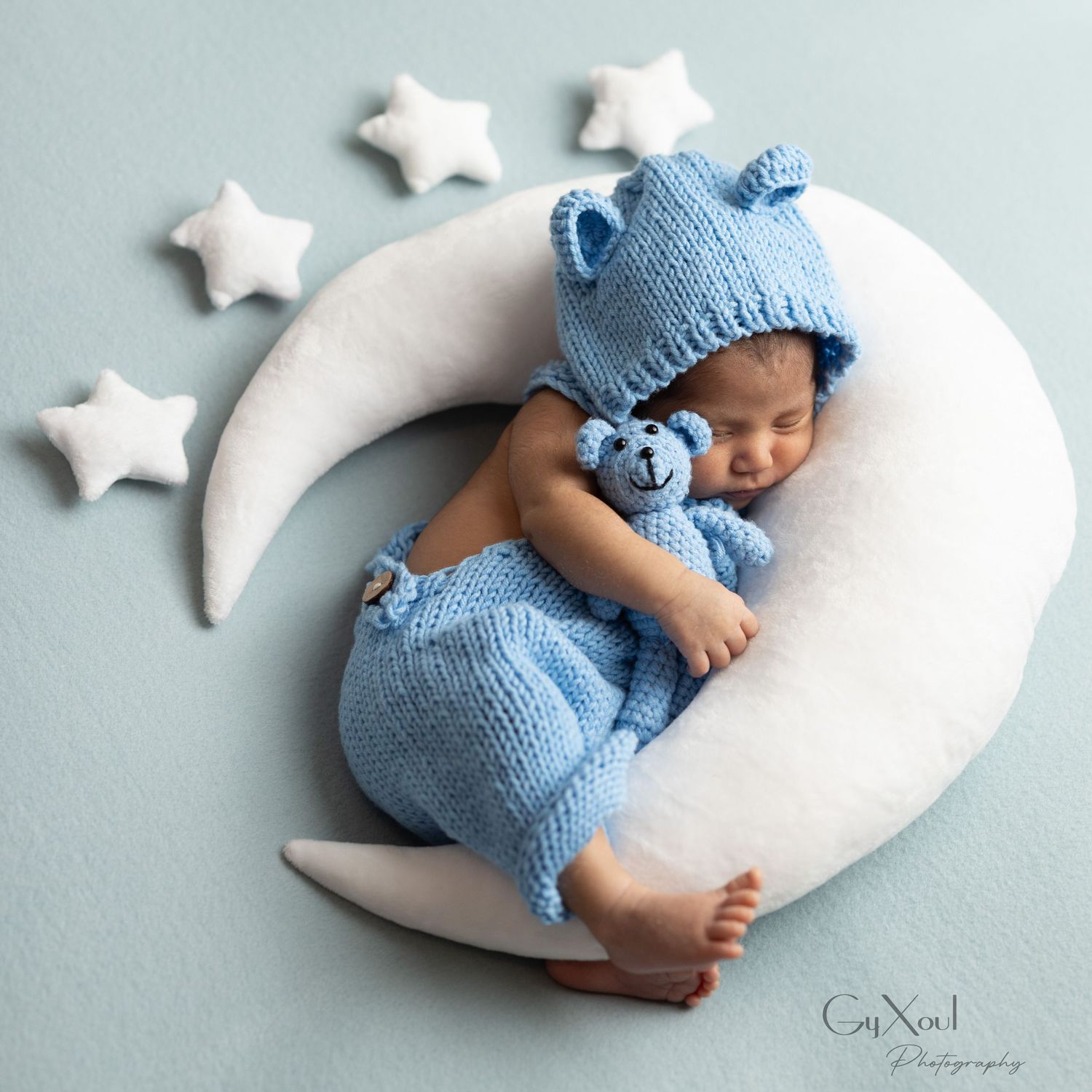 Sleeping baby in blue knitted outfit lying on decorative crescent moon cushion with star decorations on light background.