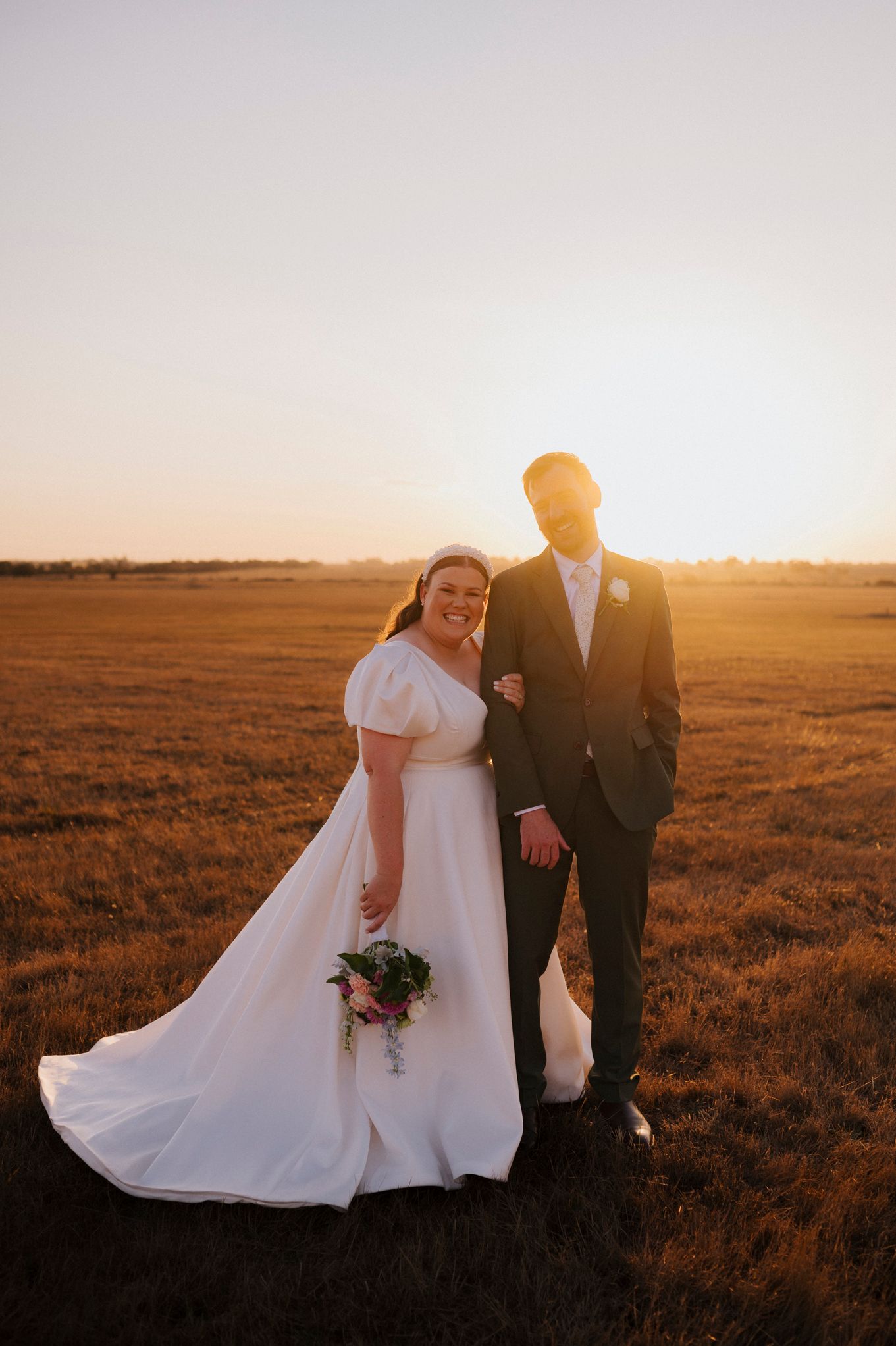 A couple in formal wedding attire poses in a field during golden hour sunset at their outdoor ceremony.