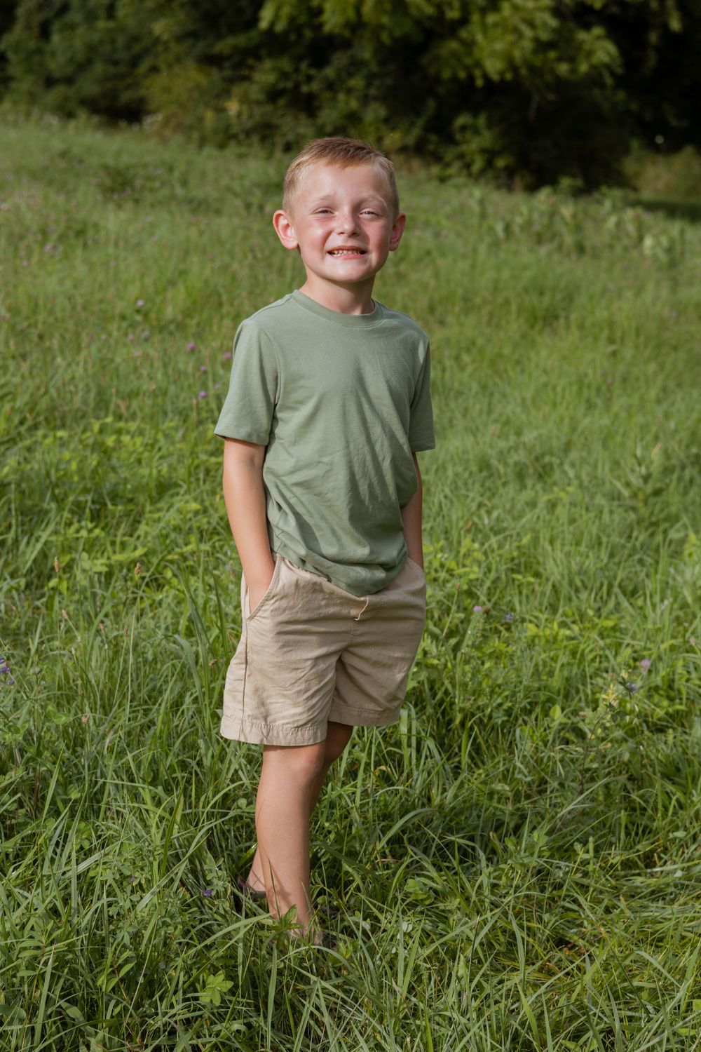 A young person in a green t-shirt and khaki shorts poses playfully in a grassy field on a sunny day.