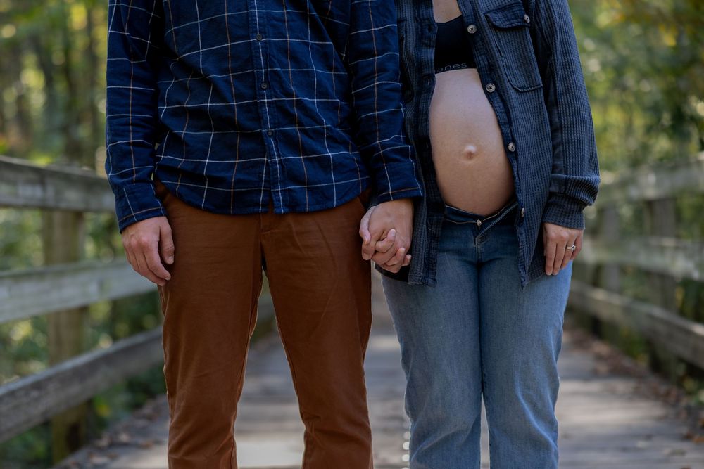 Close-up of couple holding hands on wooden bridge with pregnant belly visible.