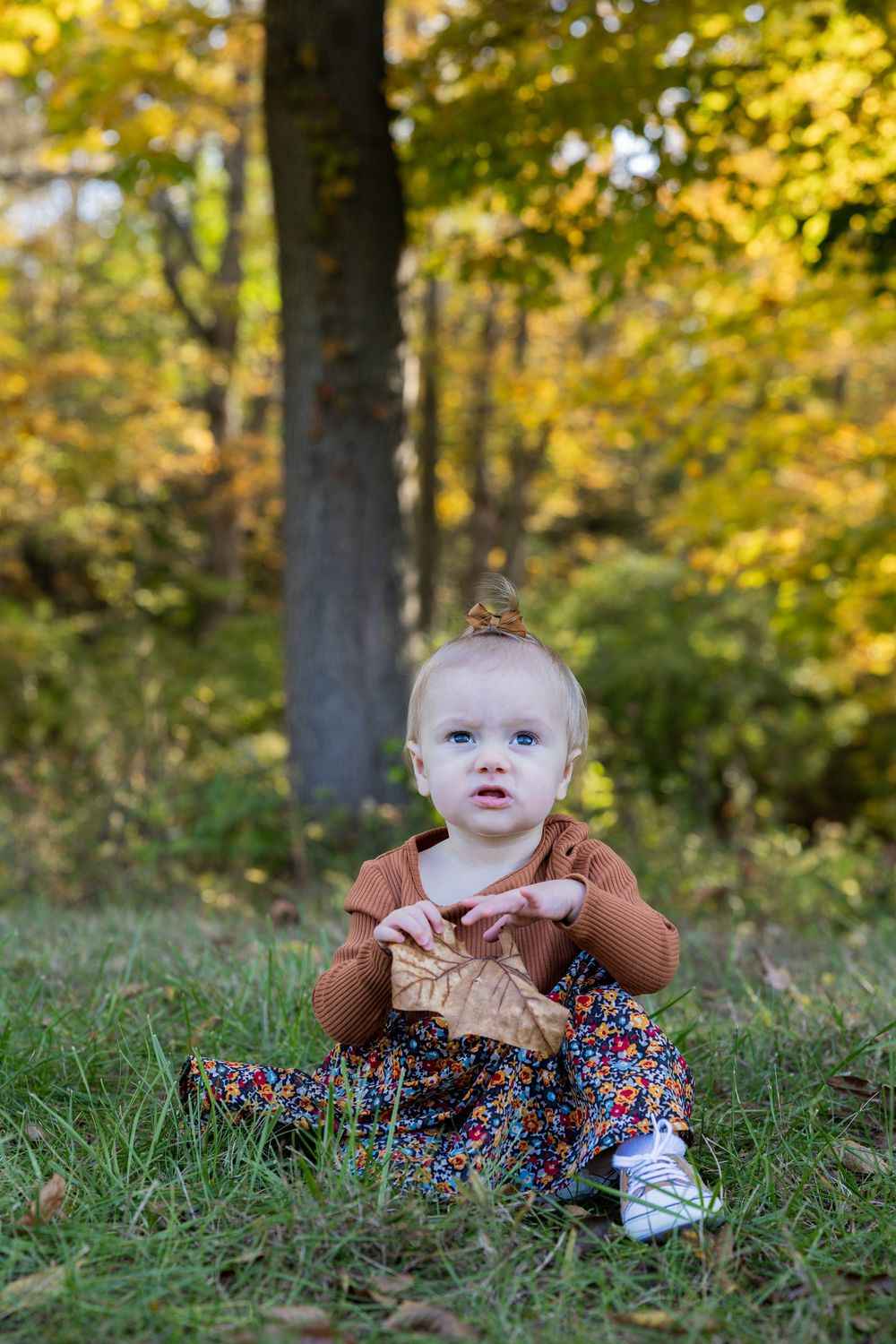 Baby in brown top and floral dress lying on grass during autumn photoshoot.