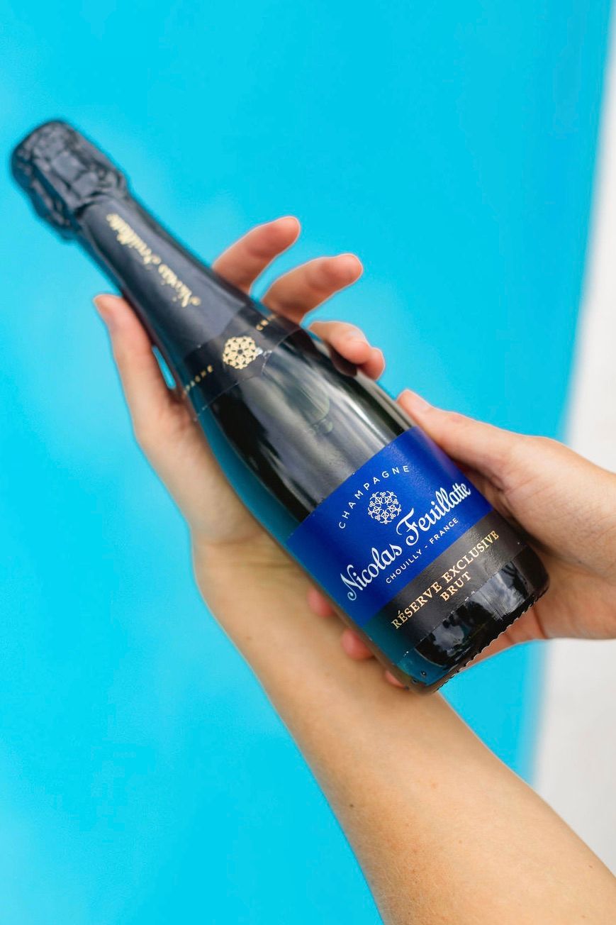 Hands holding a dark blue bottle of Nicolas Feuillatte champagne against a bright turquoise background.