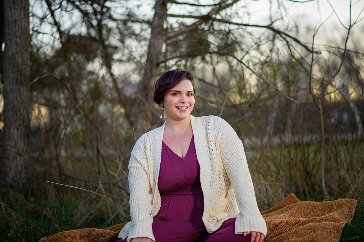 A woman in a burgundy dress and cream cardigan sits outdoors during golden hour.