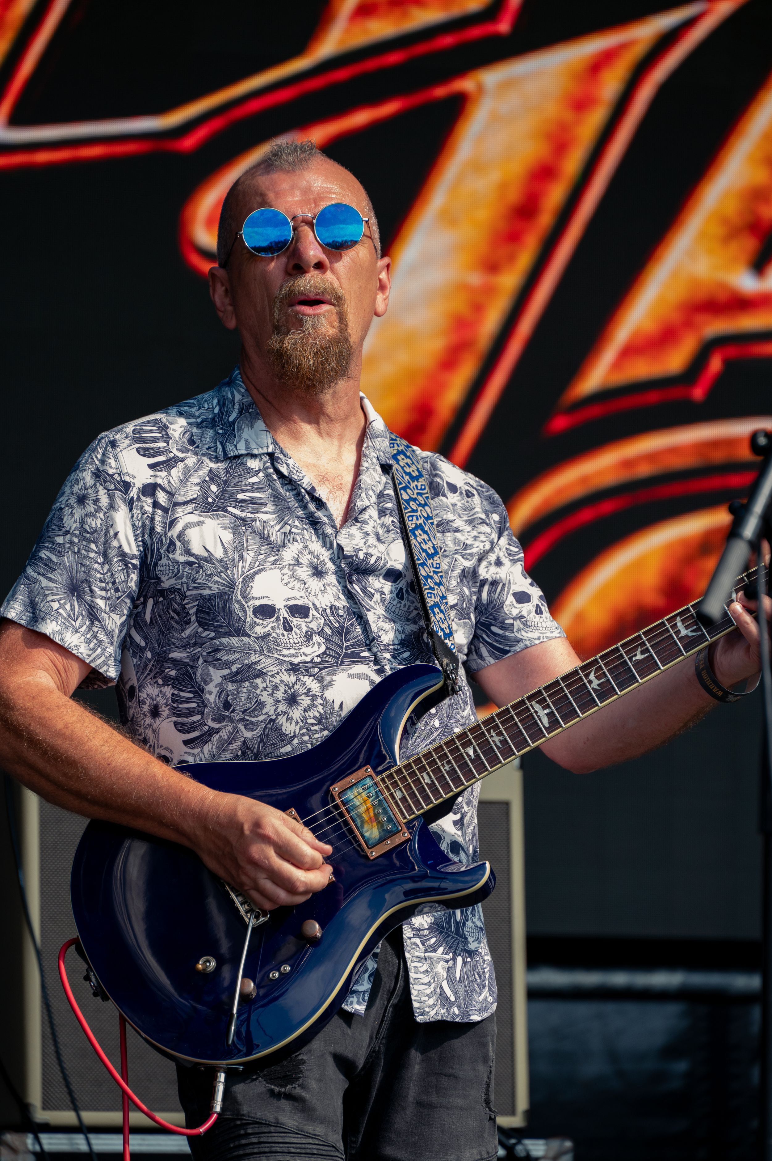 Guitarist wearing skull-print shirt performs with blue electric guitar against fiery backdrop.