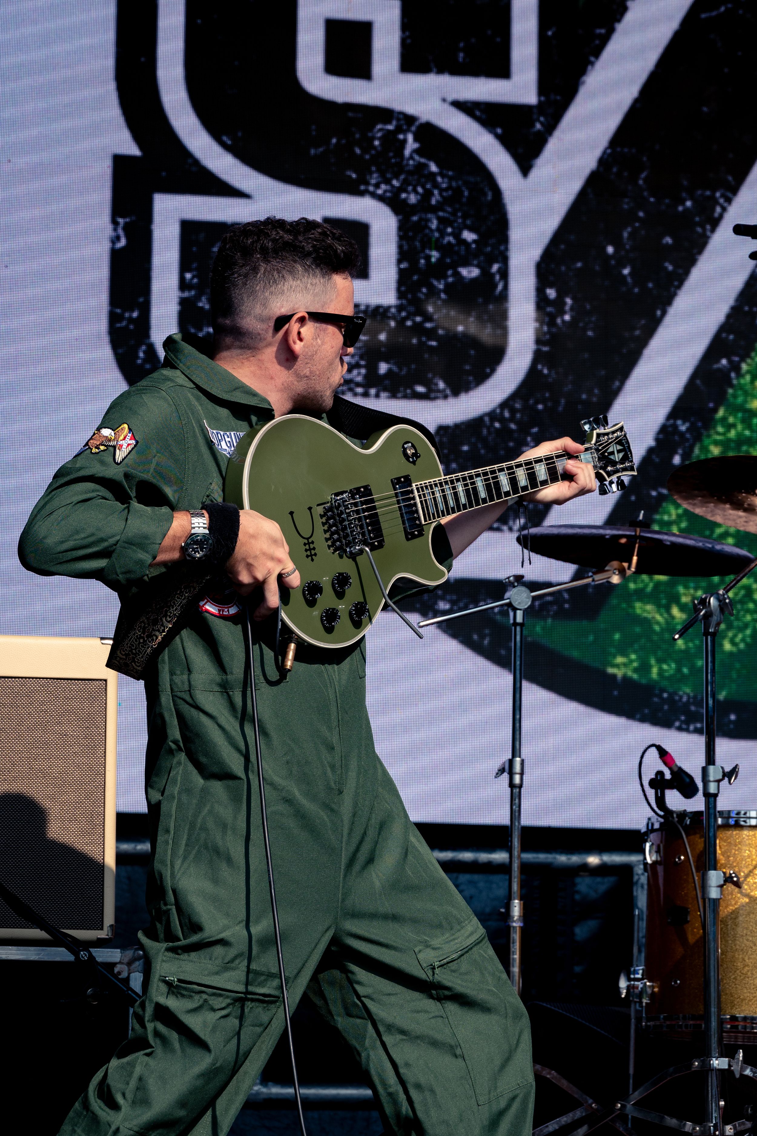 Guitarist in green jumpsuit performs with a Les Paul guitar against digital backdrop.