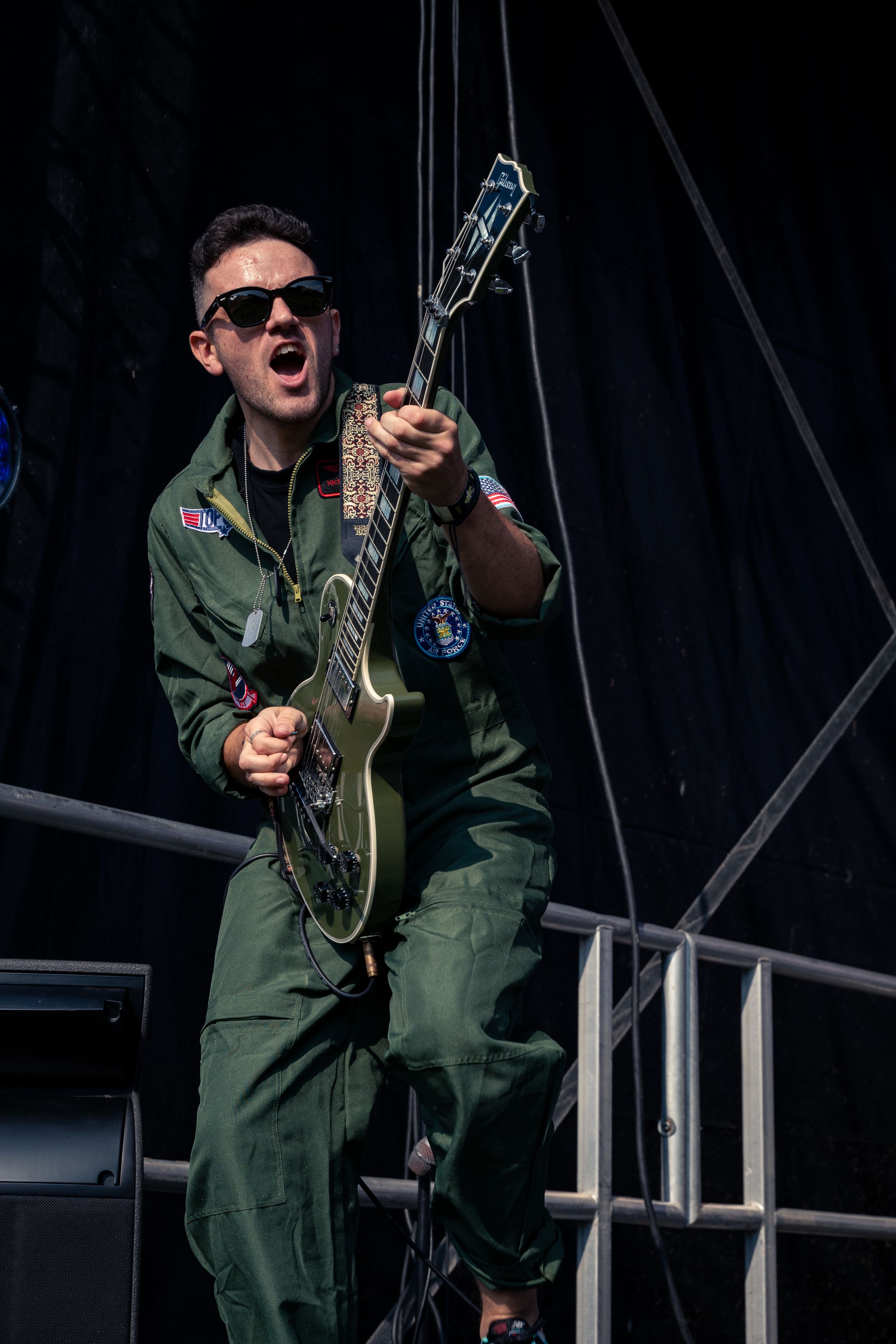 Performer in military-style jumpsuit plays green guitar energetically on outdoor concert stage.