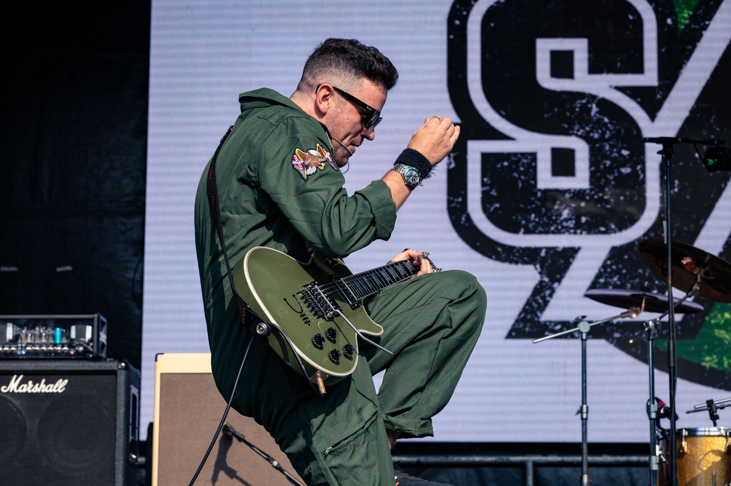 Performer in flight suit crouches while playing guitar during energetic stage performance.