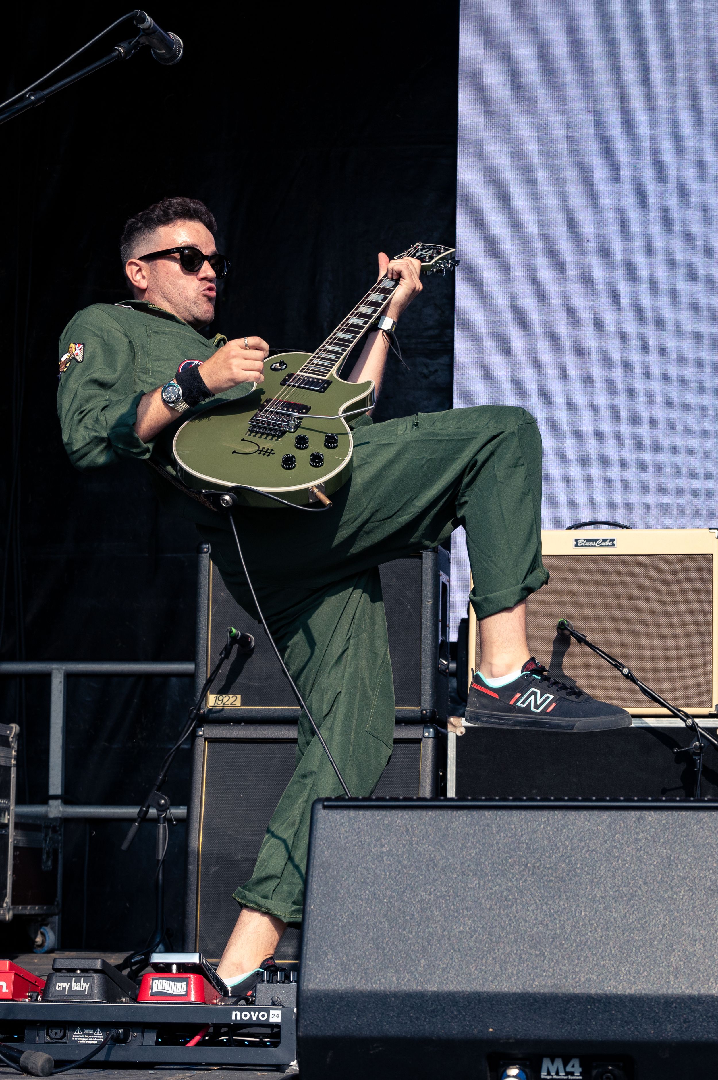 Musician in green jumpsuit strikes dynamic pose while playing guitar on elevated stage platform.