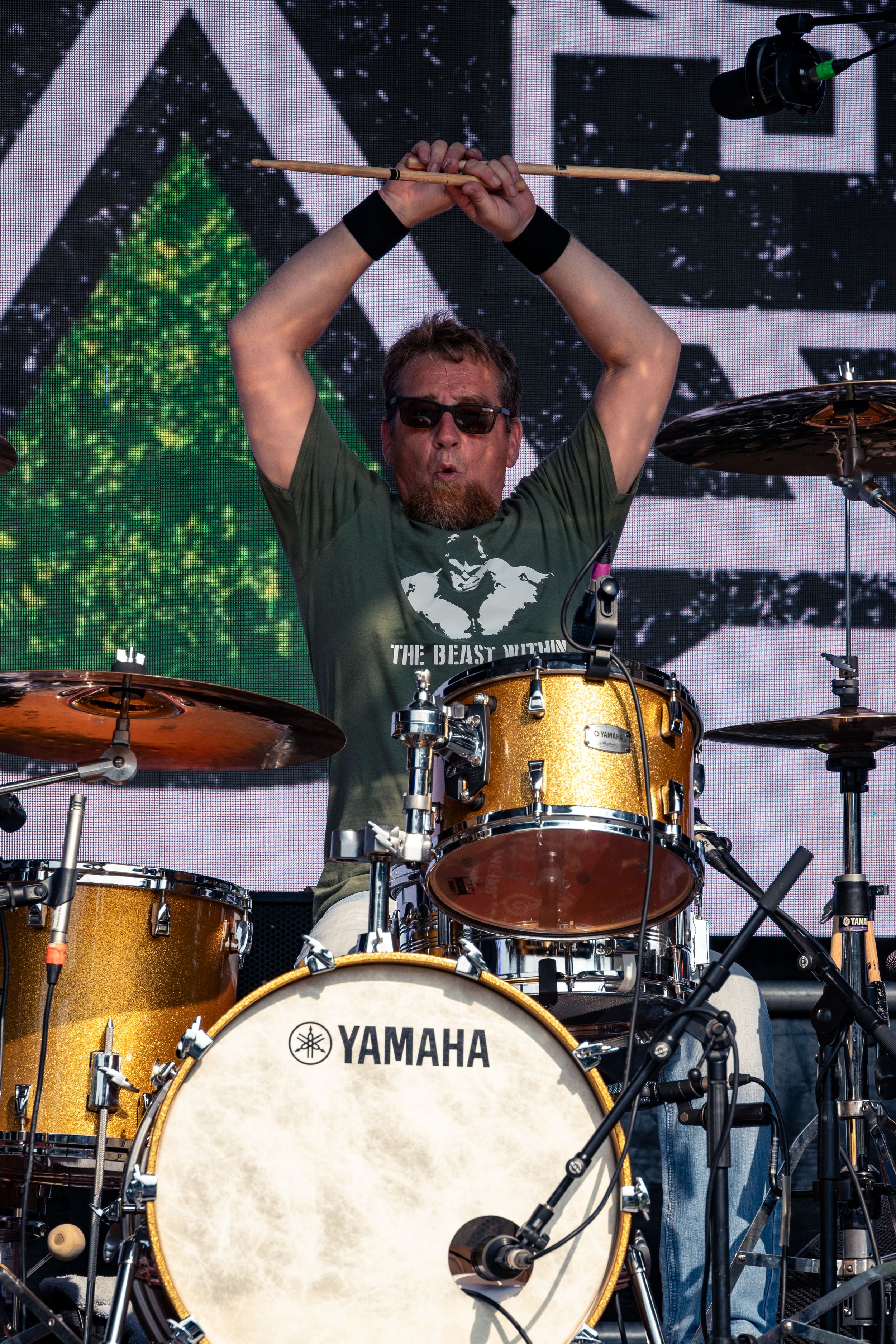 Drummer raises arms during performance behind Yamaha drum kit on festival stage.