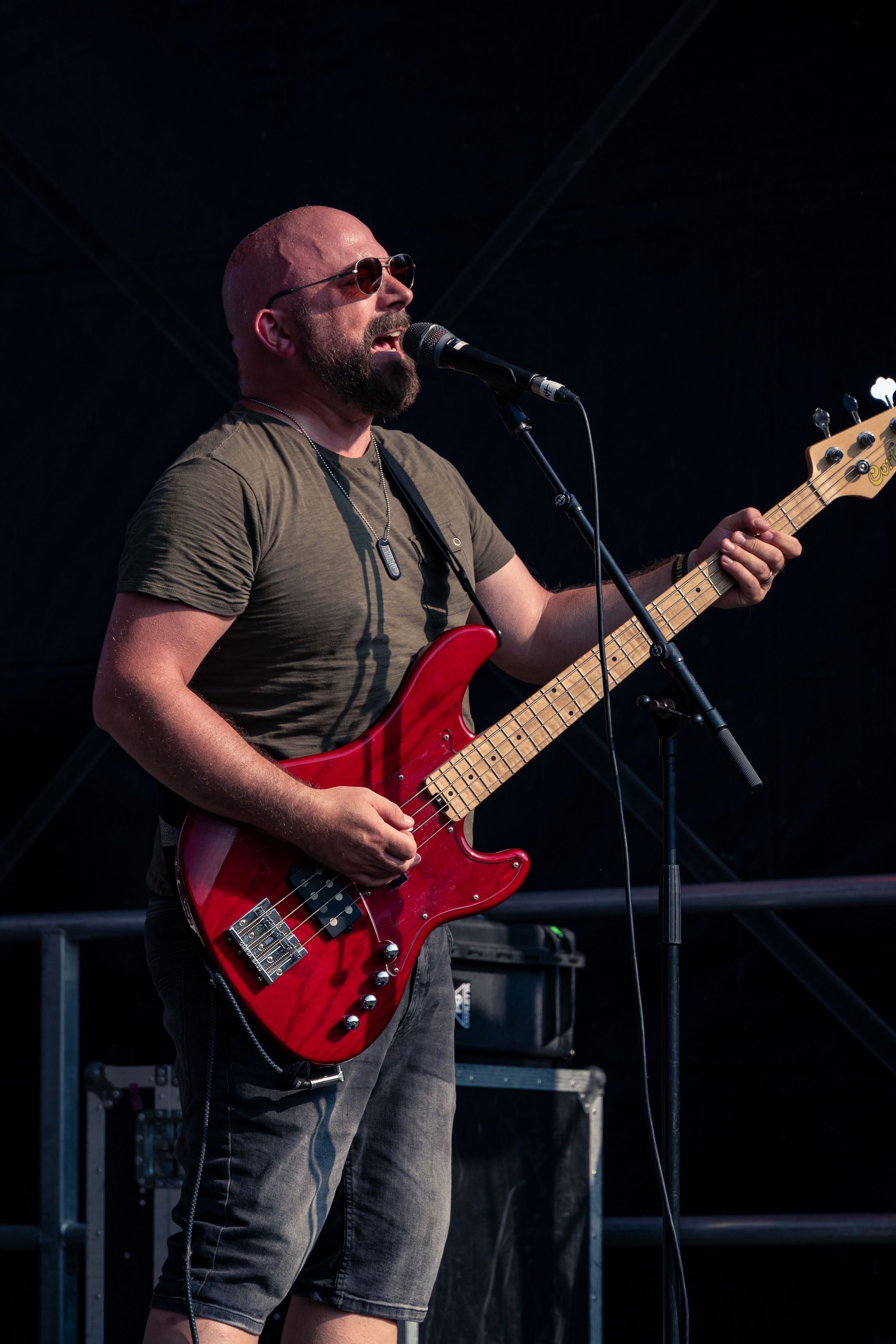 Bass guitarist in grey t-shirt and sunglasses performs on stage with red bass guitar.