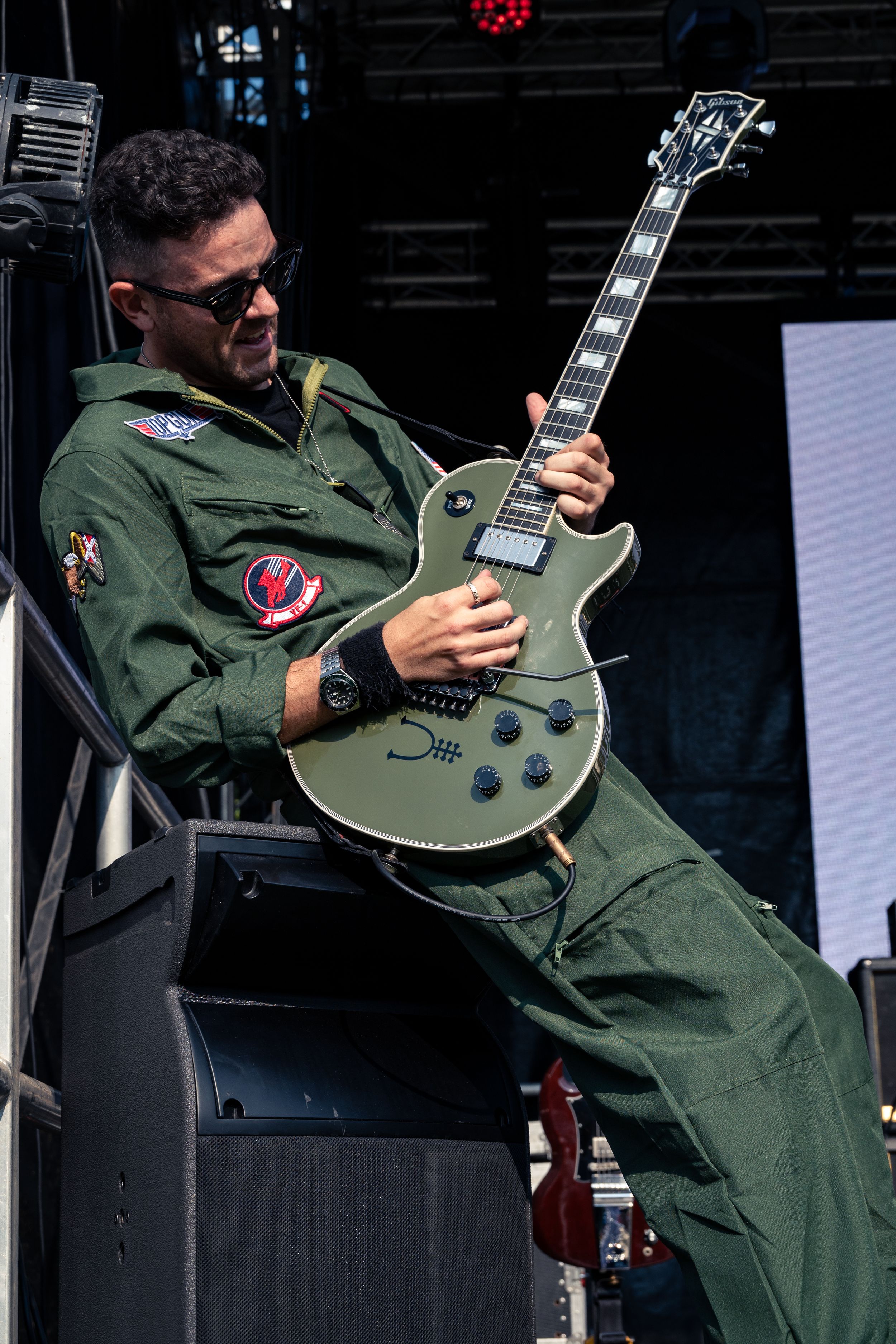 Guitarist in military-style jumpsuit plays olive green Les Paul guitar on stage.