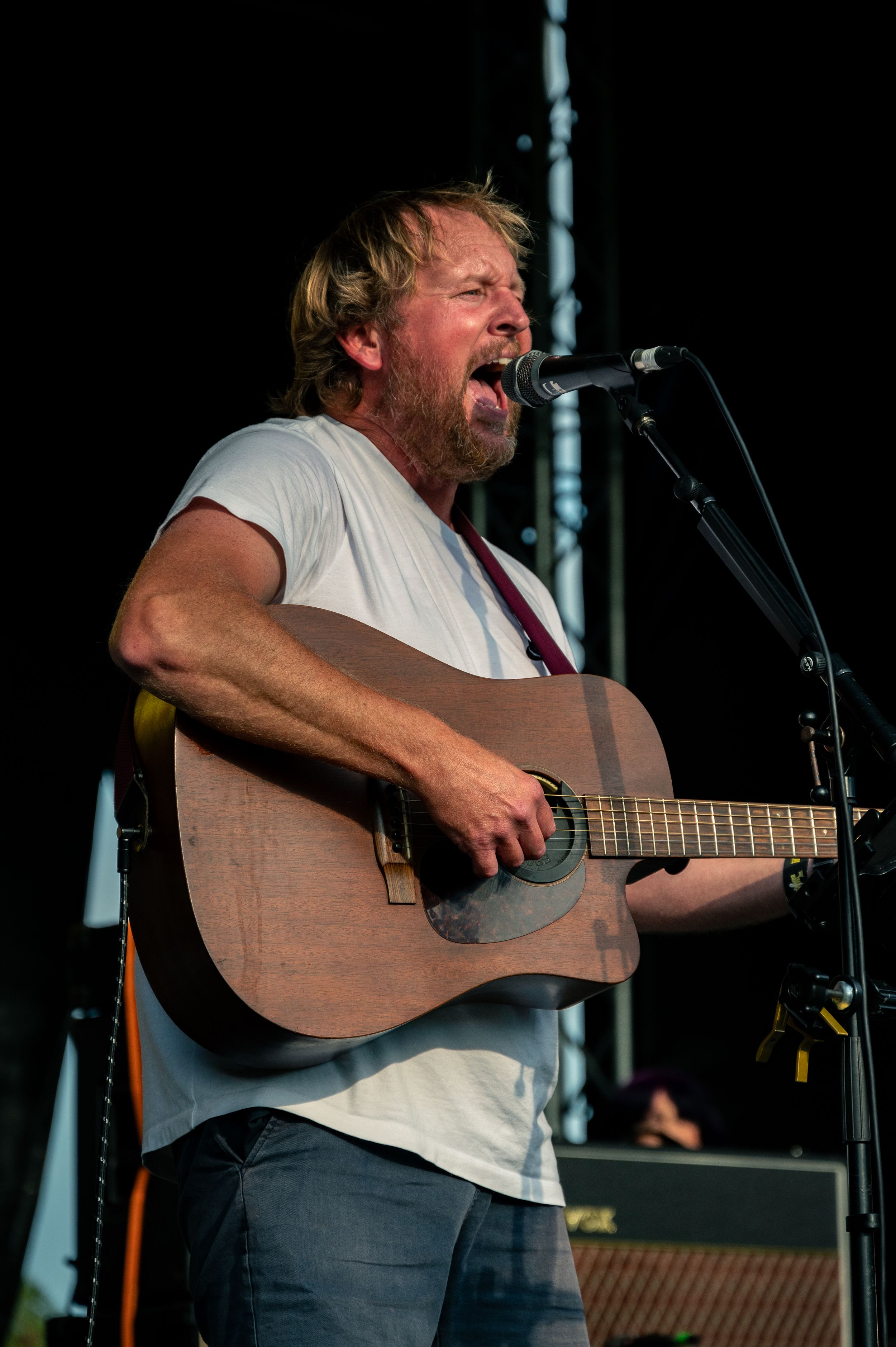 Musician sings with emotion while playing acoustic guitar on outdoor stage.