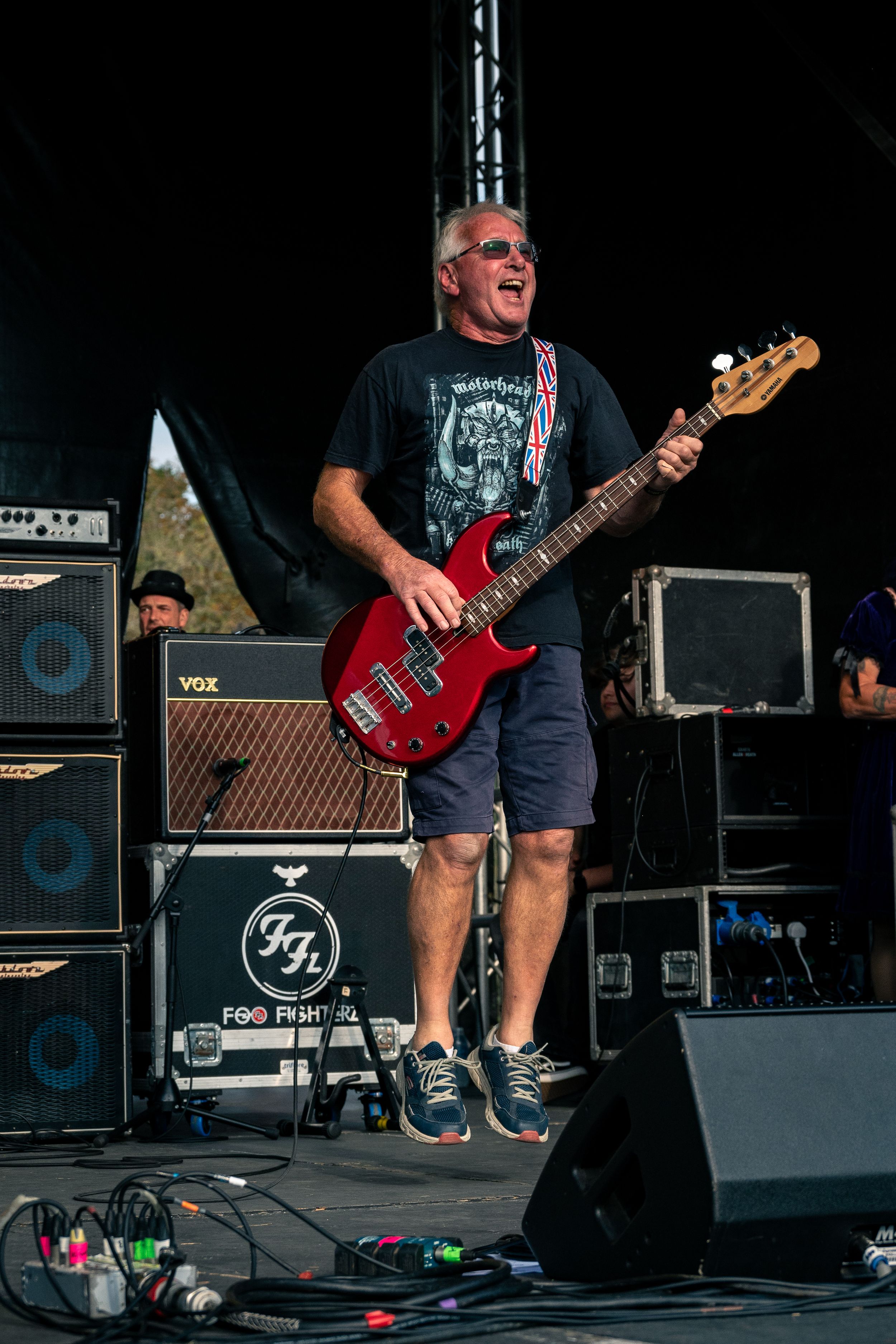 Energetic musician plays red bass guitar on stage with Vox amplifier stacks in background during outdoor concert.