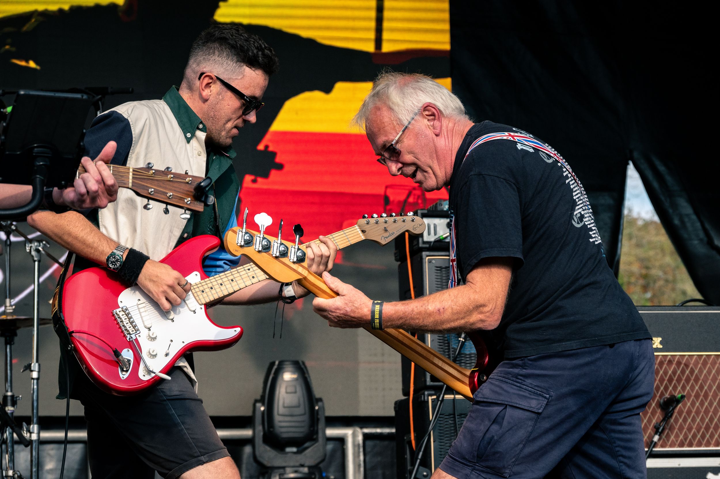 Two guitarists perform together on stage with colorful backdrop during daytime festival show.