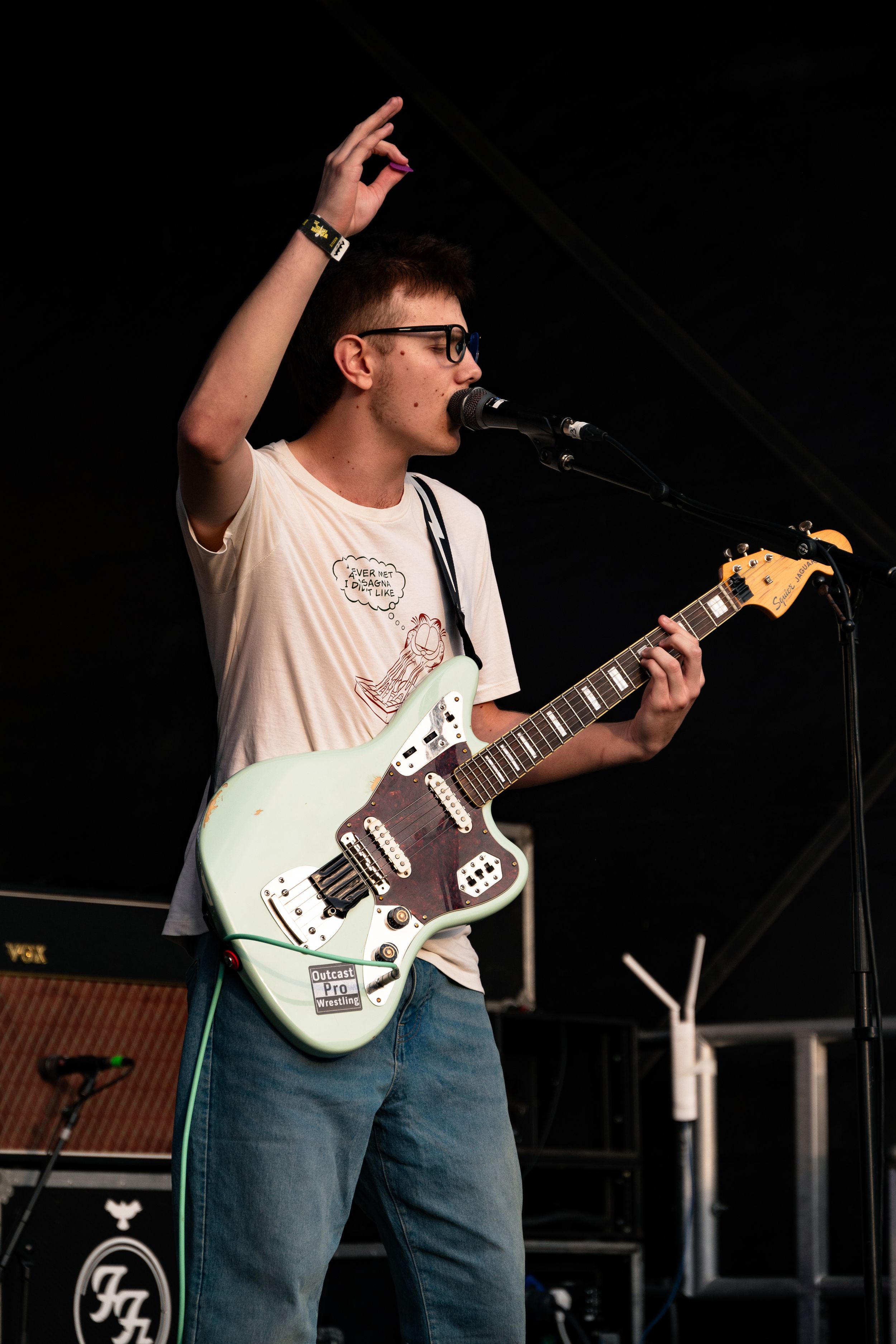 Musician in white t-shirt plays mint green guitar while singing and raising hand during performance.