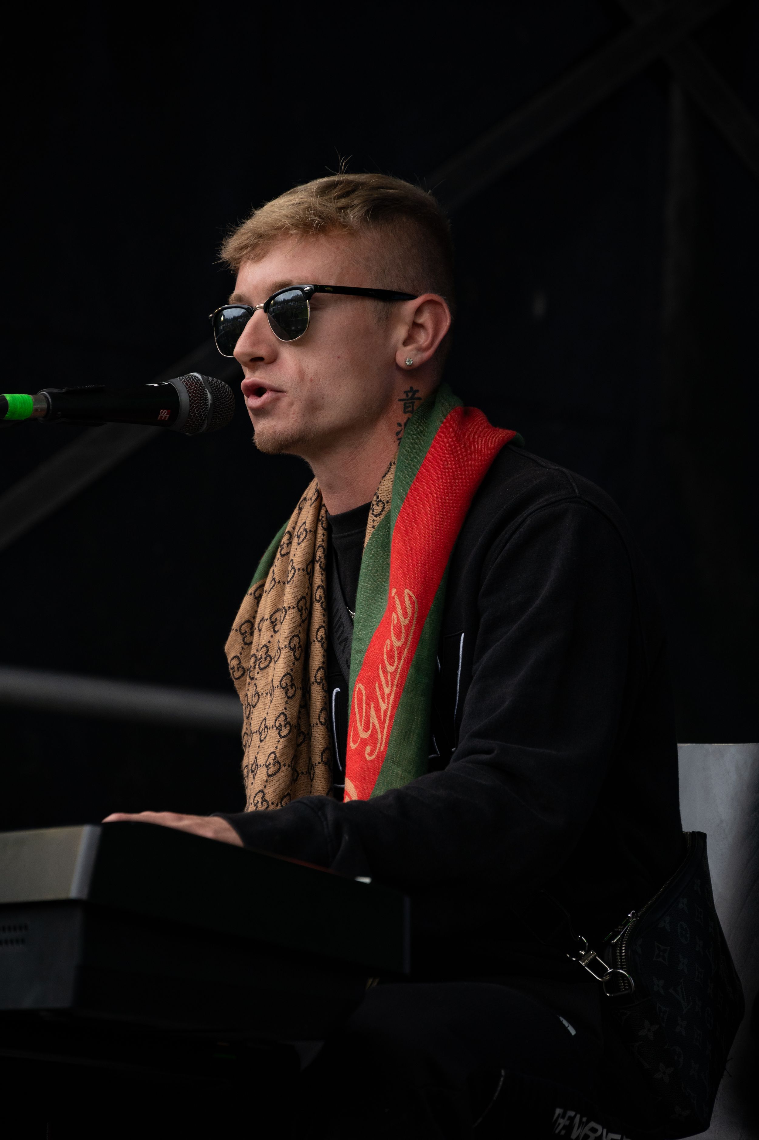 Performer in sunglasses and black hoodie sings at microphone with red guitar strap.