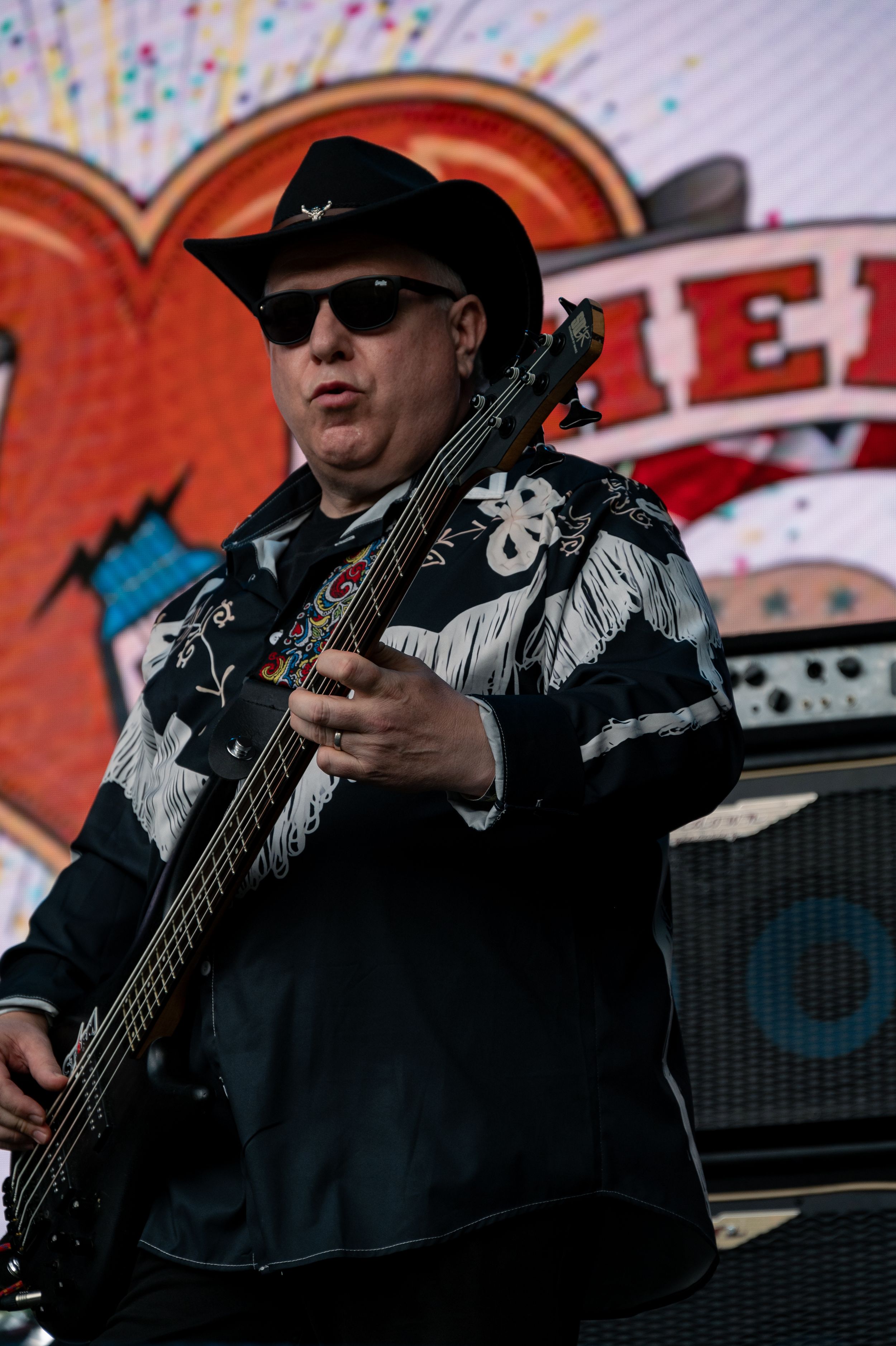 Bass player wearing cowboy hat and embroidered jacket performs on stage with colorful backdrop.