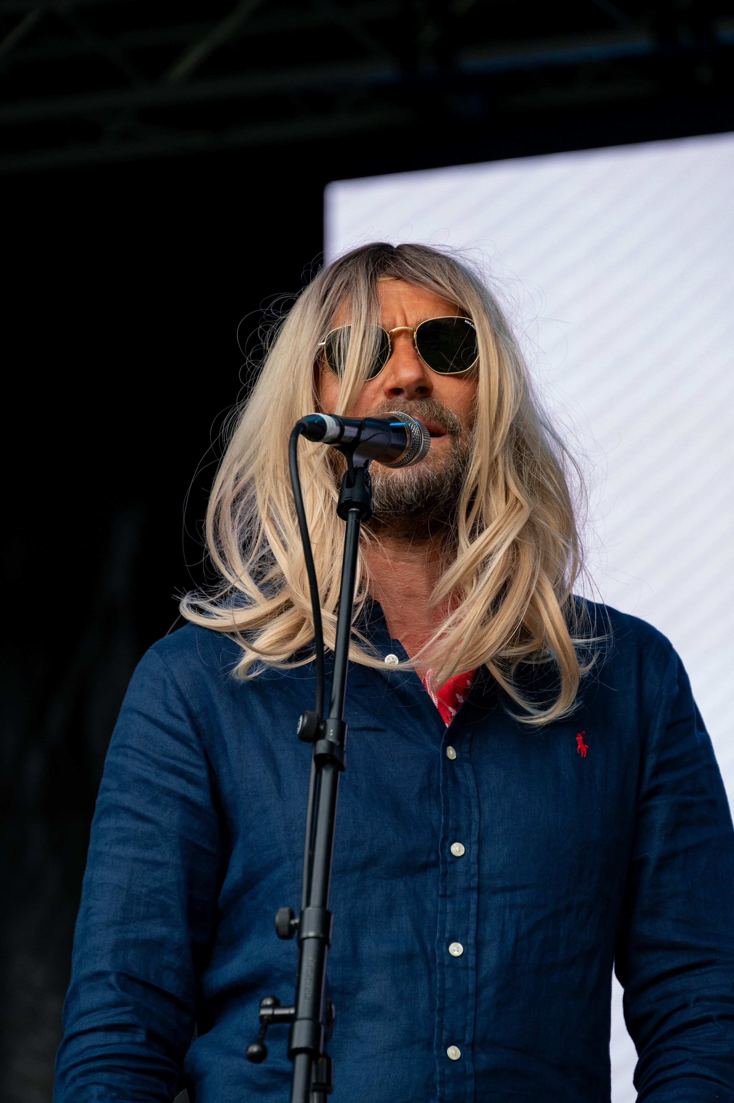 Musician with long blonde hair sings into microphone while wearing denim button-up shirt.