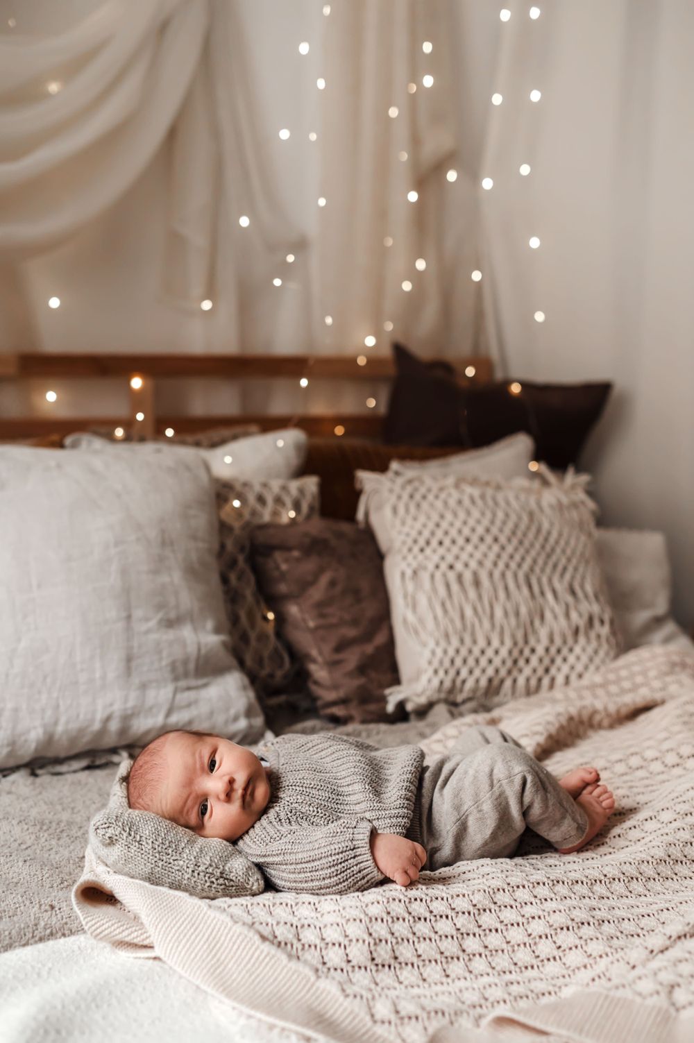 Baby in cozy sweater rests on textured bedding with twinkling fairy lights in background.
