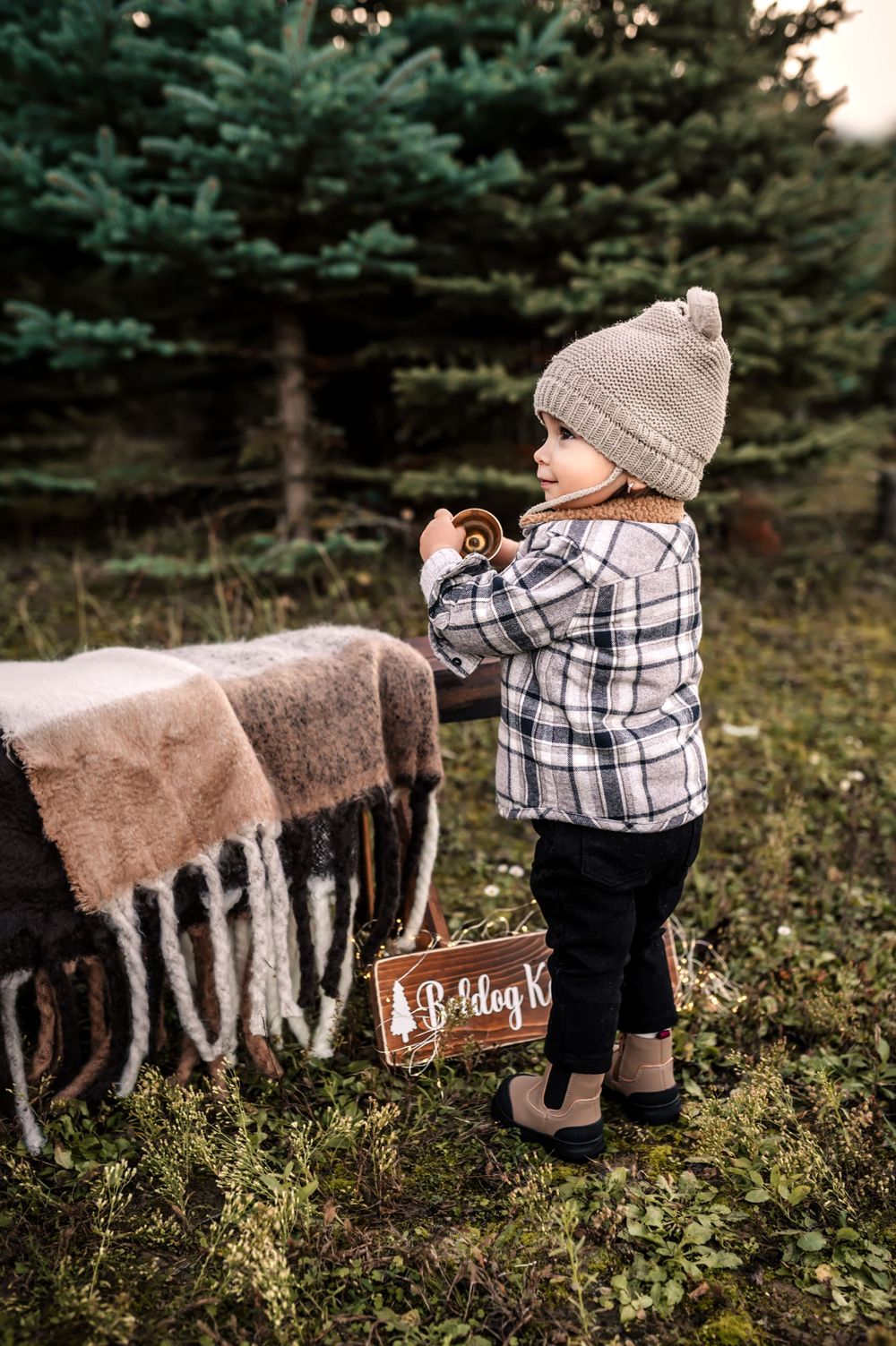 Small child in plaid shirt and knit hat stands near a cozy wool blanket in an evergreen forest setting.