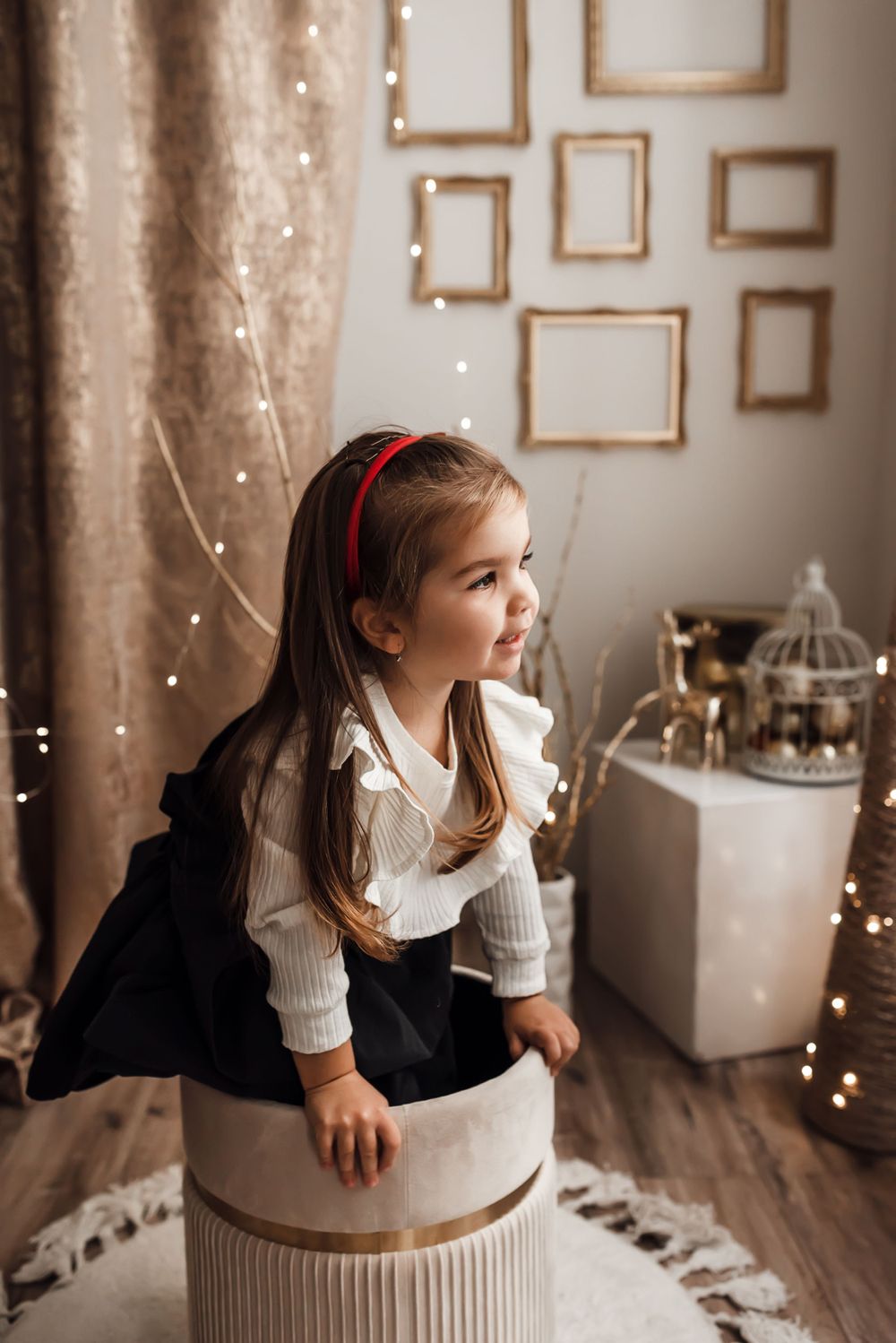 Child in elegant attire sits on a round ottoman near fairy lights and empty gold frames on wall.