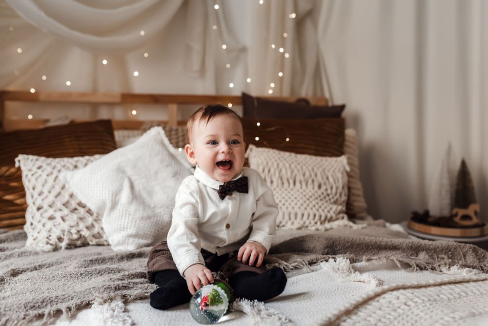 Young child in bowtie sits on textured bedding surrounded by string lights and pillows.