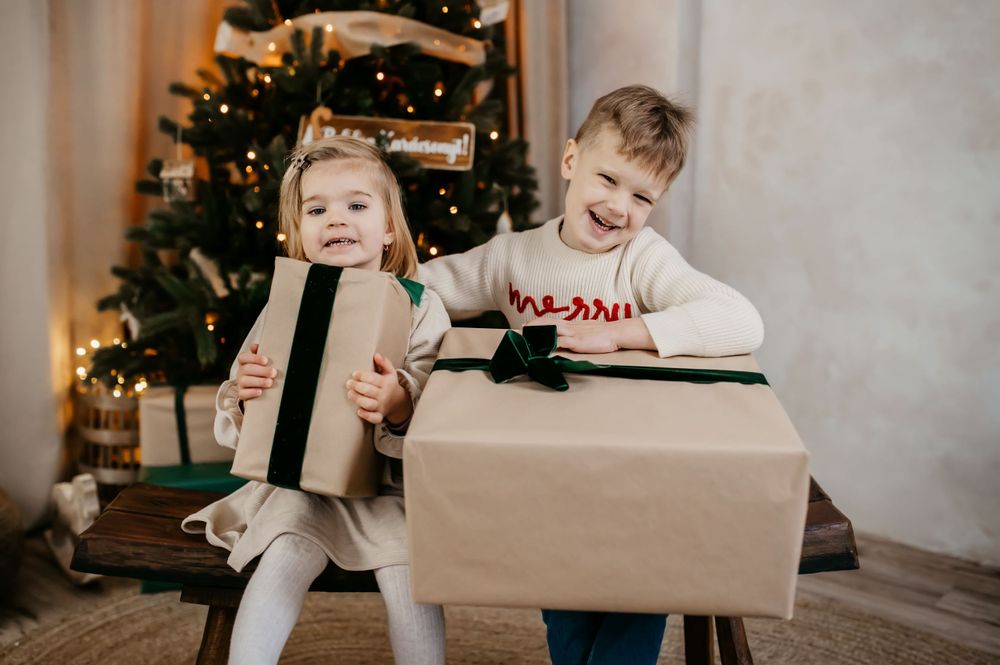 Two children laughing and playing with Christmas presents in front of a decorated tree in a cozy home setting.