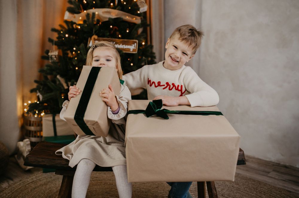 Two children excitedly unwrapping large brown gift boxes near a decorated Christmas tree with twinkling lights.