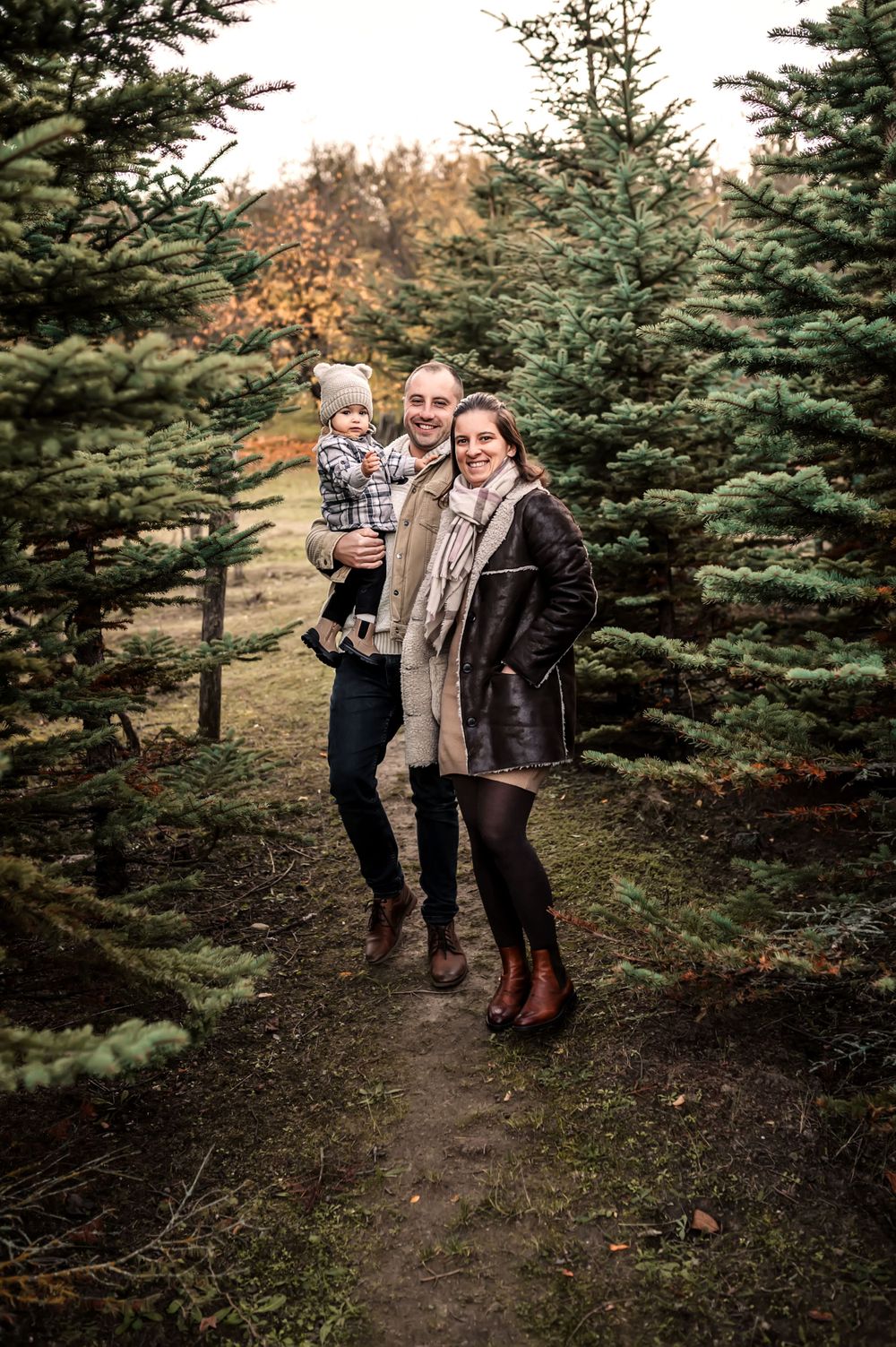 A group stands among evergreen trees at a Christmas tree farm on an autumn day with brown foliage in the background.