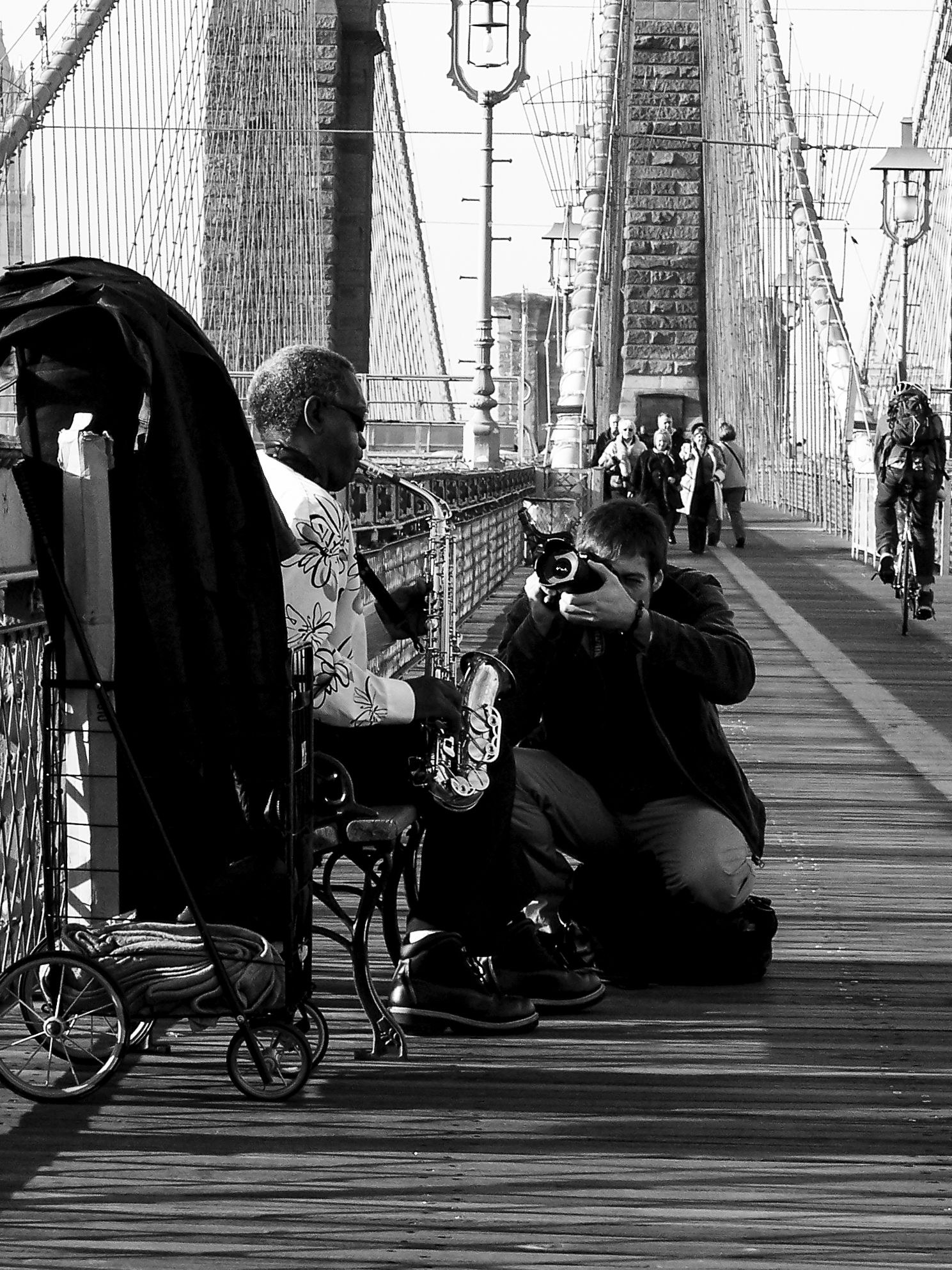 Street musicians perform with instruments on the Brooklyn Bridge walkway while pedestrians pass by in black and white.