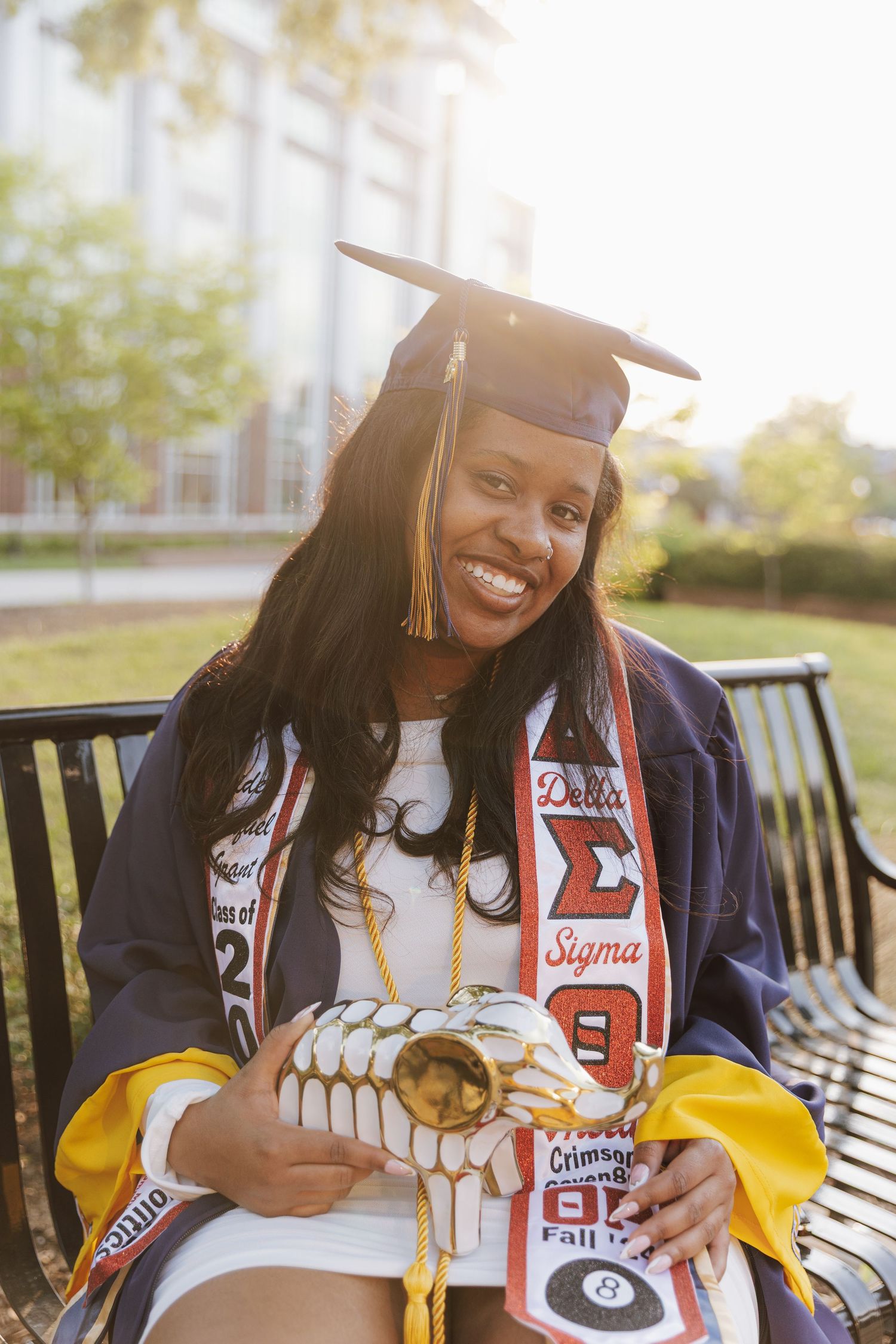 Graduate wearing navy blue academic regalia and white polka dot dress poses near university entrance sign on sunny day.