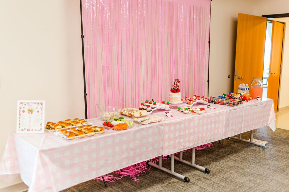 Long buffet table with pink backdrop and white tablecloth set up for a celebration.