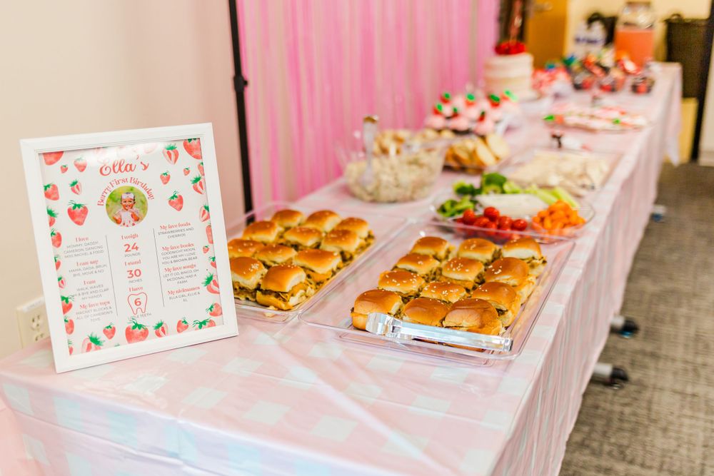 Pink themed party buffet table with slider sandwiches and a menu sign featuring strawberry decorations.